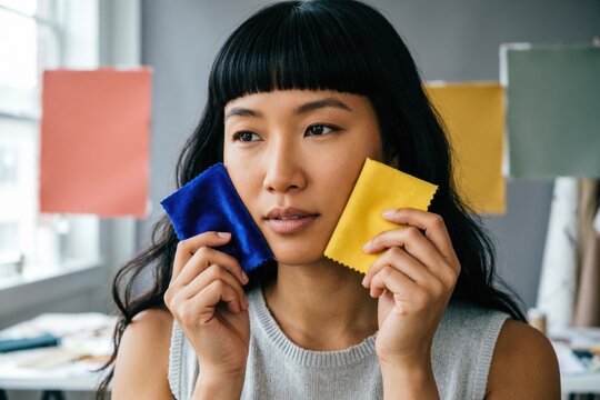Una mujer coreano-estadounidense con flequillo recto, sosteniendo muestras de tela o de color cerca de su rostro