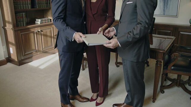 Passing gray binder from right man to men in law-office, woman in burgundy suit guiding review