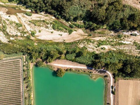 Aerial view of Laghi Santarelli featuring a vibrant turquoise lake surrounded by lush green trees, agricultural fields, and a winding dirt path in Moresco, Marche, Italy.