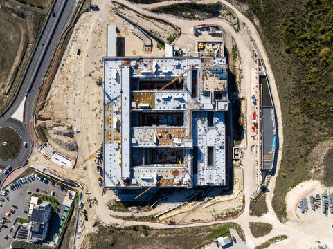 Aerial view of an Hospital large construction site featuring a multi-wing building structure with cranes, surrounding dirt grounds, and nearby road infrastructure in Ancona, Marche, Italy.