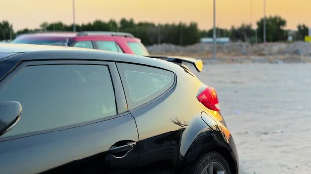 A glossy dark hatchback car is parked in an outdoor dirt lot during a warm sunset. The evening light reflects off the shiny exterior paint, tinted windows, and rear spoiler.