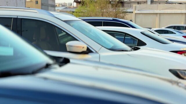 A row of modern vehicles sits parked in an outdoor urban lot during bright daylight. A shallow depth of field highlights a white SUV while blurring the foreground car and background building.