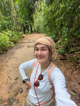 Smiling young Asian woman takes a selfie on a dirt path in a lush forest.