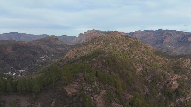 An aerial drone shot pushes in on the rugged, pine-covered mountain landscape of Gran Canaria. The iconic Roque Nublo dominates the horizon under an overcast sky, with a remote village in the valley.