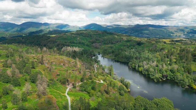 Calm river winds through green valley. Clouds drift above distant mountain peaks. Tarn Hows is a stunning, accessible beauty spot in the Lake District National Park in Cumbria, England