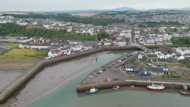 Coastal town nestles beside calm harbor waters. Boats rest quietly at pier, surrounded by low-lying buildings. Grey sky looms overhead. Maryports located in the heart of the Lake District in Cumbria