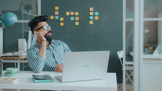 Arab businessman in casual clothing and glasses is talking on mobile phone and using laptop typing at work enjoying modern business and technology.