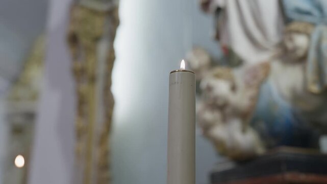 A lit candle burns on a chapel altar, with a painted statue and gilded carving out of focus around it.