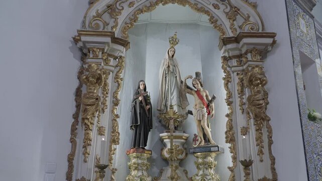 A gilded baroque altar niche with statues of Our Lady of Fatima, a nun holding a crucifix and Saint Sebastian, beside an azulejo tile panel.