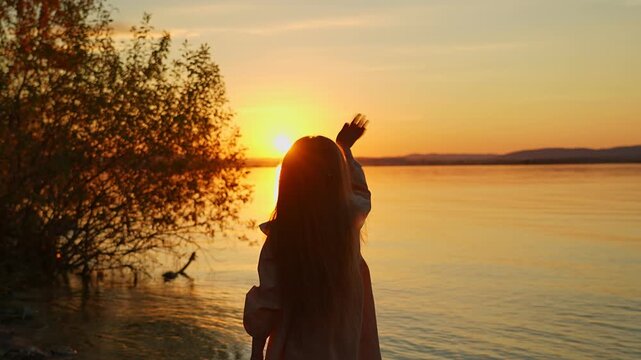 A little girl is standing on the shore of a lake at sunset and joyfully waving her arms at the golden sun. A moment of childlike delight and inspiration.