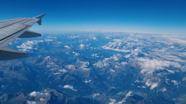 Aerial perspective through an airplane window reveals the sunlit South Tyrolean Alps below, with a small section of the wing in view and wisps of clouds floating around the mountain peaks