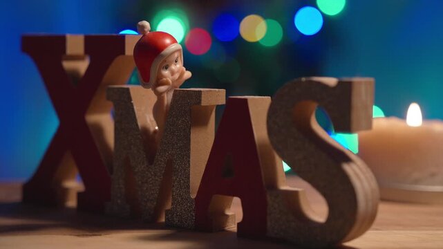 A small Santa Claus figurine looks out from among wooden XMAS letters as a candle glows softly beside colorful Christmas tree lights