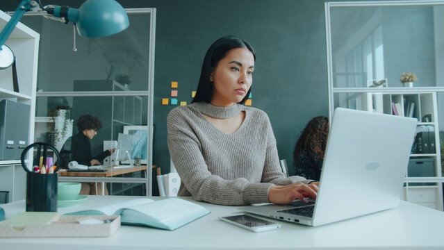 Mixed race businesswoman is using laptop then massaging temples feeling headache in workplace, women are working in background. People and health concept.