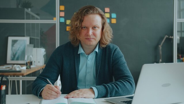 Portrait of attractive young man looking at camera with serious face holding pen sitting at desk in modern office. People, career and paperwork concept.