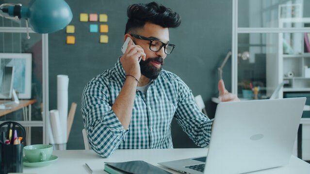 young Middle Eastern man speaking on mobile phone in office sitting at desk talking and gesturing. Communication and job concept.