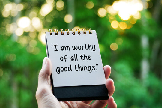 Hand holding notebook with text "I am worthy of all the good things." in nature.