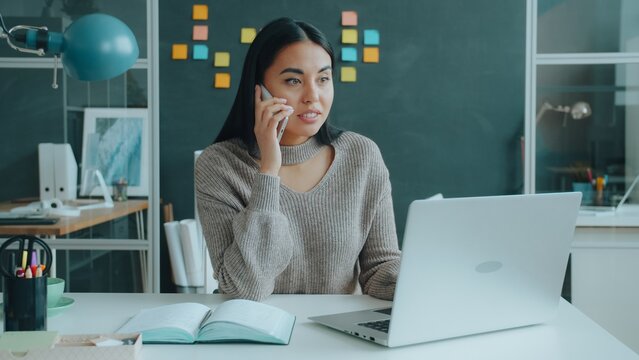 Happy mixed race woman is talking on mobile phone getting good news using laptop typing in office at desk. Business, success and emotions concept.