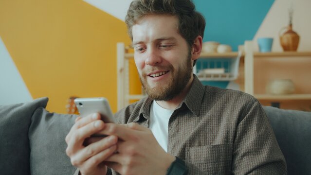 handsome guy using smartphone texting touching screen relaxing on couch in modern apartment. Devices and communication concept.