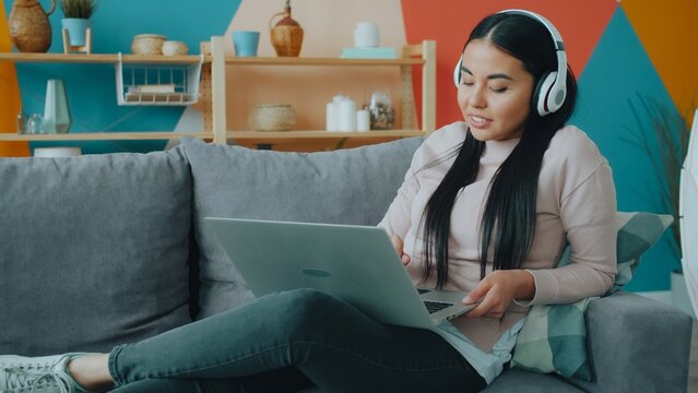 Attractive Asian girl wearing headphones is using laptop computer relaxing on couch at home enjoying online content. Leisure time, youth and devices concept.