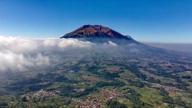 Aerial view of Mount Merbabu rising above the clouds with lush green terraced fields and villages in the foreground in Selo, Central Java, Indonesia.