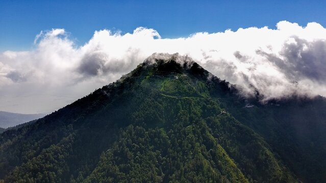 Aerial view of Mount Telomoyo covered in dense green forest with clouds drifting over the peak in Getasan, Central Java, Indonesia.