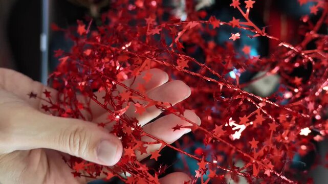 Close-up video of a hand touching and arranging shiny red Christmas tinsel decorated with small star shapes, with blue ornaments visible in the background