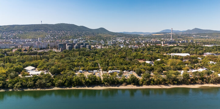 Aerial view of the wooded Danube shoreline and festival grounds on Obudai-sziget, with Buda residential districts and the Buda Hills behind, in Budapest, Hungary.