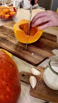 a person uses a knife to cut a piece of orange pumpkin on a wooden board, garlic cloves and a glass jar sit on a nearby surface
