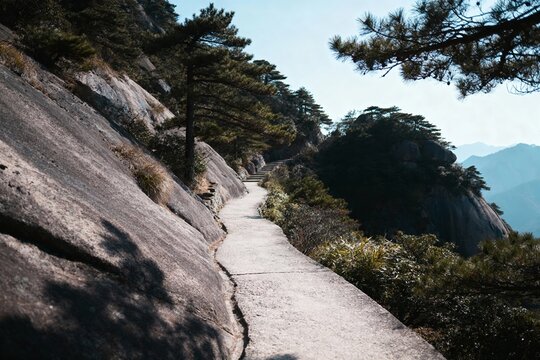 A winding stone path ascends through a mountainous landscape with rocky cliffs, pine trees, and distant hills under a clear sky.