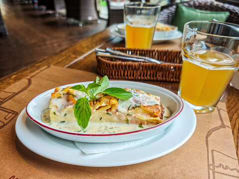 Portion of lasagne or stuffed pasta rolls served with a creamy sauce, garnished with a sprig of basil, along with glasses of drink