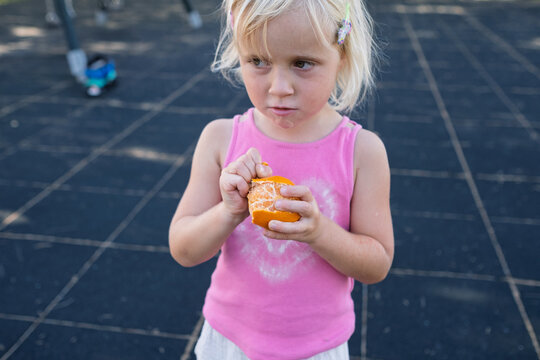 Cute Little Blonde Girl Peeling Fresh Tangerine at Outdoor Playground
