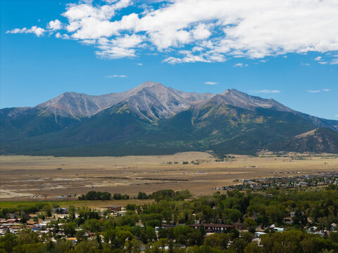 Aerial view of the Collegiate Peaks mountain range rising above the town of Buena Vista with green trees and open plains under a bright blue sky in Buena Vista, Colorado, United States.