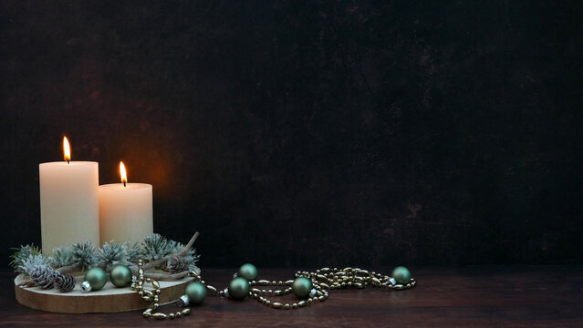 Zwei brennende wei&szlig;e Kerzen auf Holzscheibe mit Tannenzweigen, Zapfen und gr&uuml;nen Weihnachtskugeln auf dunklem Hintergrund mit Textfreiraum.