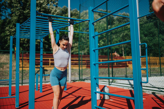 Fit young woman performs a hanging pull-up on outdoor monkey bars while wearing workout gloves. Athlete trains strength and balance on blue bars at a sunny outdoor fitness park.