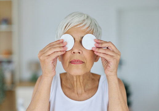 Portrait of a senior elderly woman cleaning face with a cotton pad and her skin in front of mirror in the bathroom in the morning