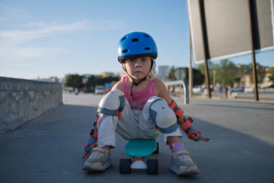 Little Girl in Blue Helmet and Protective Gear Sitting on Penny Board at Skatepark