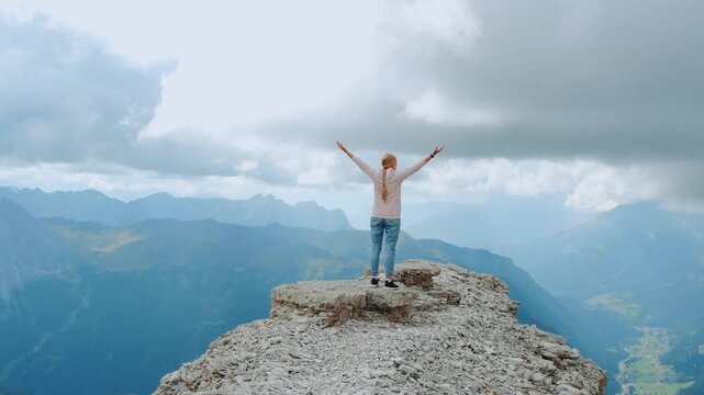 A young blonde woman with outstretched arms stands on a large rock in a vast mountainous area under an overcast sky during daytime.