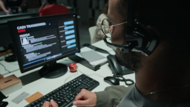 Biracial young man wearing headset typing on keyboard and monitoring cash transfer on computer screen. Suitable for cybersecurity training and scam awareness videos