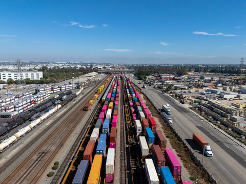 Aerial view of a busy railyard filled with colorful shipping containers on freight trains, adjacent to industrial roads and warehouses in Carson, California, United States.