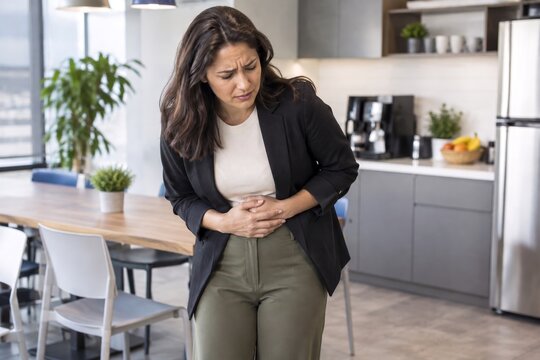 Young Professional Woman Clutching Her Stomach In Pain While Standing In Office Breakroom, Depicting Digestive Issues Or Gastritis