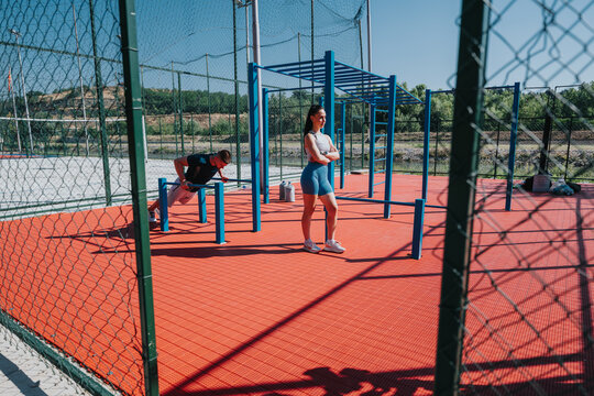 A woman stands at an outdoor calisthenics park while a man performs dips on parallel bars. The scene shows outdoor fitness training with workout equipment and athletic activity.