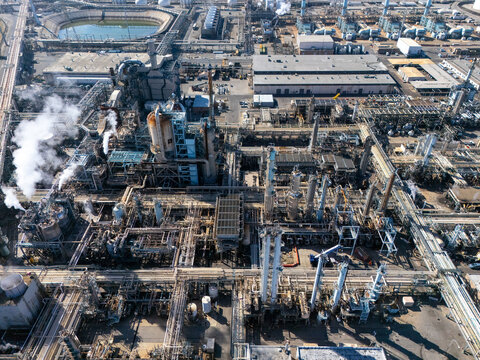 Aerial view of an industrial oil refinery in Carson, featuring complex piping, storage tanks, tall distillation towers, and smoke rising from a vent, Carson, California, United States.