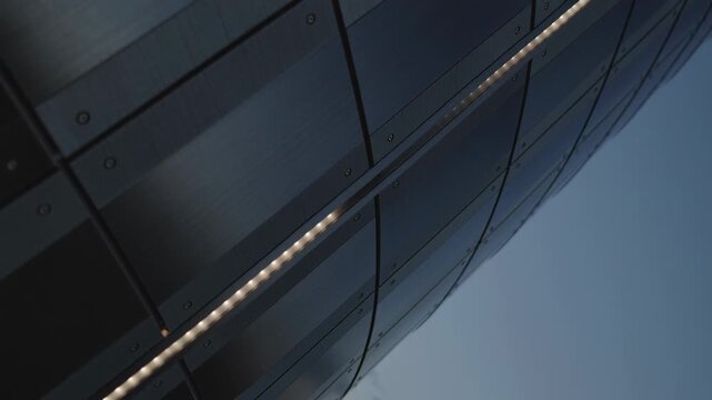 Tracking curved riveted metal facade dusk as small orange glow appearing and moving along LED seam