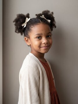Pensive girl with hair bows standing sideways against neutral wall in soft light