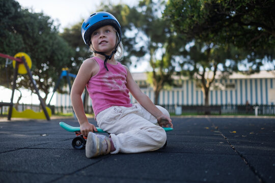 Cute Little Girl in Blue Helmet Sitting on Penny Board at Playground Outdoors