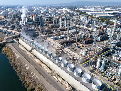 Aerial view of the oil refinery complex with cooling towers, industrial pipes, and storage tanks under a clear sky in Carson, California, United States.