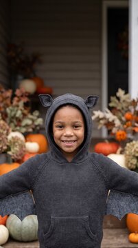 Mischievous child in bat hoodie costume smiling on porch decorated with pumpkins and dried flowers
