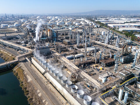 Aerial view of a large oil refinery complex featuring numerous storage tanks, cooling towers emitting steam, and complex piping networks in Carson, California, United States.