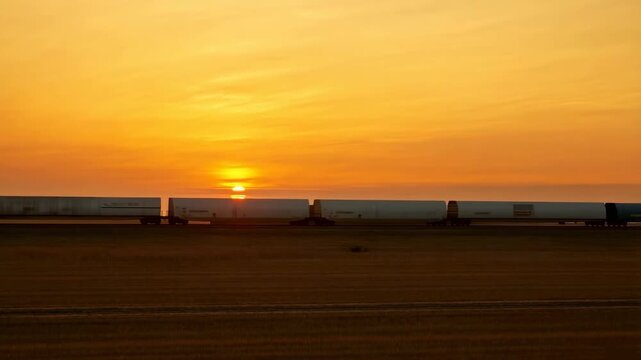 Freight train moves across open farmland beneath a glowing orange sunset