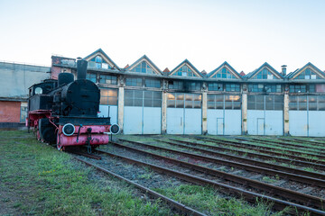Curved facade of an old locomotive roundhouse near Dej, Romania, with multiple gabled bays and steam engine in the foreground, showing industrial railway heritage. © Anze